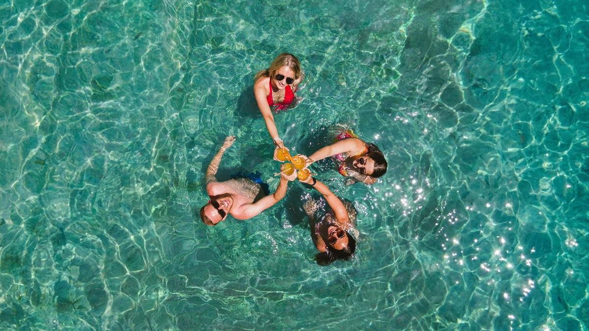 Overhead frame of four guests clinking orange cocktails in a turquoise pool